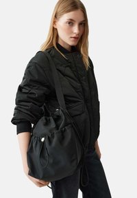 Giovane donna con capelli biondi che indossa un giubbotto imbottito nero e tiene una grande borsa a tracolla nera con cordino su uno sfondo semplice.