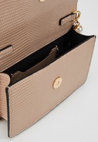 Béžová kabelka s hadím vzorem a zlatým knoflíkovým zapínáním, otevřená a odhalující černý interiér a malou kapsičku se zlatým a perlovým přívěskem.