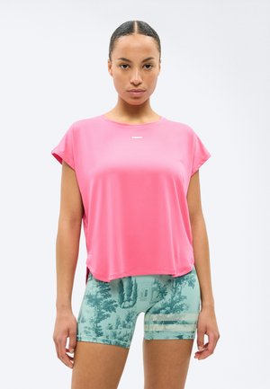 Femme portant un haut ample rose vif et un short de vélo vert à motifs, debout avec une expression neutre sur un fond uni.