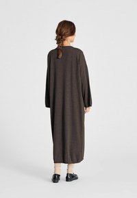 Ženska s pletenico, oblečena v dolgo temno rjavo obleko, bež nogavicami in črnimi čevlji, stoji ob enobarvni svetli podlagi.