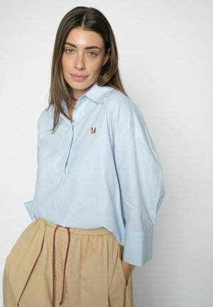 Kobieta o długich brązowych włosach, ubrana w jasnoniebieską luźną koszulę z wyhaftowaną literą M oraz beżowe spodnie z sznurkiem, stojąca na białym tle.