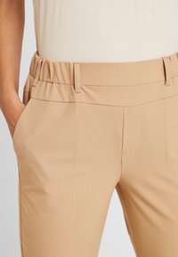 Pantalons beiges ajustés avec une taille élastique, des poches avant et un tissu lisse et extensible pour le confort et l'ajustement.