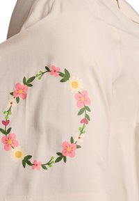 Lysebeige bluse med blomsterdesign med pink blomster, grønne blade og hvide accenter, placeret midt på ærmet. Blød tekstur.