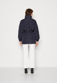 Námořnická pufovaná bunda s vysokým límcem, páskem v pase a vodorovnými švy. Modelka má na sobě bílé široké kalhoty a černé boty.