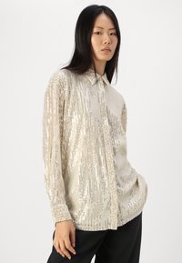 Blusa scintillante in paillettes argentate con colletto sartoriale e chiusura a bottoni, caratterizzata da una vestibilità ampia e strisce verticali texturizzate.