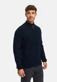 Suéter de punto acanalado azul marino con cremallera y cuello alto, con tirador marrón en contraste, que presenta mangas largas y un corte holgado.