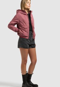 Donna in piedi con le mani in tasca, indossa una giacca con cappuccio rosa polvere, una maglietta nera, pantaloncini di jeans scuri e stivaletti neri, su uno sfondo semplice.