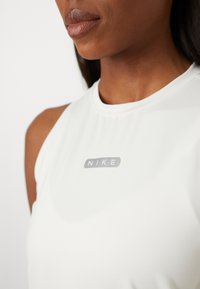 Vit ärmlös atletisk topp med en slät yta, som har en grå "NIKE"-logotyp centrerad på bröstet. Rund halsringning och en minimal design.