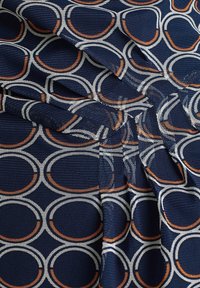 Marinfärgad tyg med överlappande cirkulära mönster i vitt och orange. Texturen är slät med ett veckat mönster som skapar visuell intress.