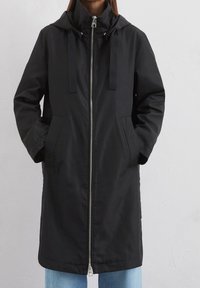 Svart vattentålig rock med dragkedja fram, huva, justerbara snoddar, framfickor och en slät textur. Mittenlång design.