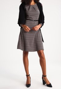 Femme portant une robe noire à motifs longueur genoux avec une ceinture, un cardigan noir drapé sur les épaules, et des talons à bride cheville noirs.