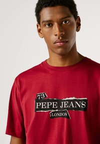 Camiseta de algodón roja con cuello redondo y mangas cortas. Presenta un estampado gráfico negro con los textos "PEPE JEANS" y "LONDON".