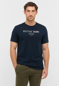 Námořnickomodré tričko vyrobené z bavlny s kulatým výstřihem. Obsahuje bílé grafické texty "MUSTANG JEANS" a má slim fit design.