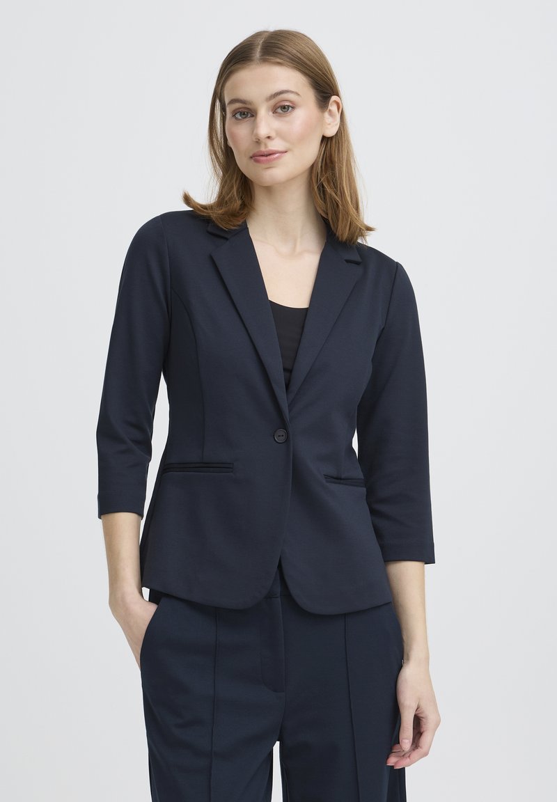 Blazer bleu marine à manches trois-quarts, fermeture à un bouton, deux poches avant, et texture lisse. Associé à un pantalon assorti.