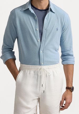 Mann trägt hellblaues Hemd mit Knöpfen über einem marineblau gestreiften T-Shirt mit hochgekrempelten Ärmeln, weiße Shorts mit Kordelzug und Armbanduhr.