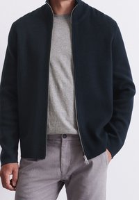 Giacca nera con zip realizzata in un tessuto a maglia, con colletto alto, abbinata a una maglietta grigia chiara a girocollo e pantaloni grigi chiari.