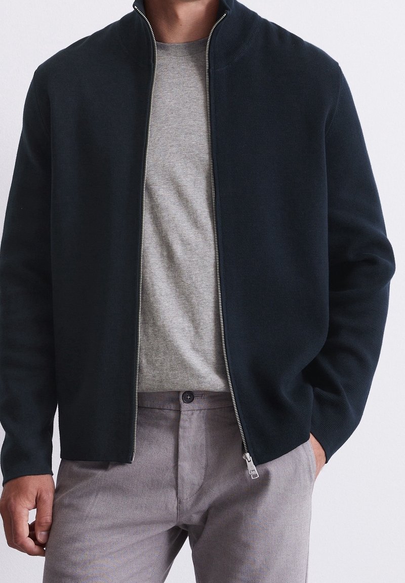 Giacca nera con zip realizzata in un tessuto a maglia, con colletto alto, abbinata a una maglietta grigia chiara a girocollo e pantaloni grigi chiari.
