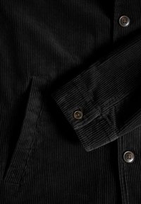 Black corduroy jacket featuring ribbed texture, button closures, and a side pocket. Detail of collar and sleeves shown.