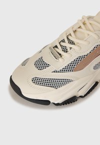 Sneakers beige avec des accents crème et des panneaux en mesh noir, bout rond, semelle épaisse, et lacets crème sur un fond blanc.