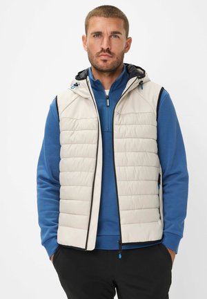 Lichte crème puffer vest met een capuchon, voorzien van een ritssluiting aan de voorkant, zwarte afwerking en blauwe accenten, gedragen over een blauwe langslijfs top.