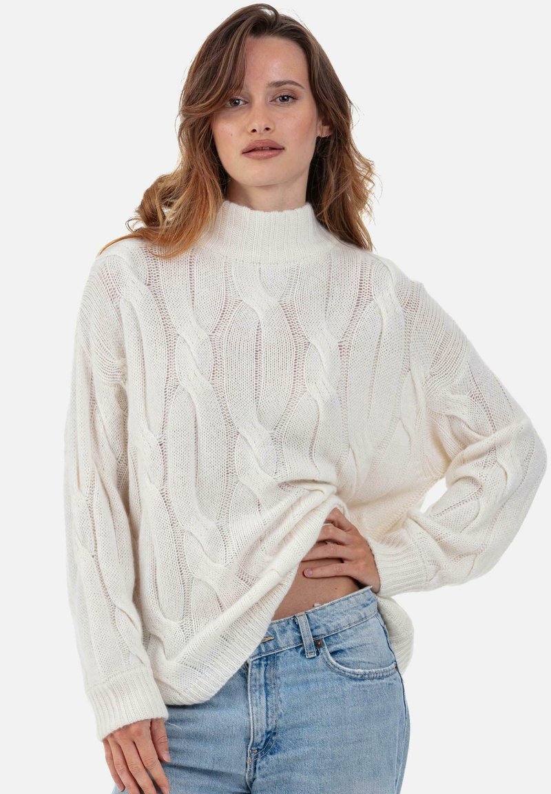 Cremefarbener Strickpullover mit Zopfmuster, hohem Kragen, langen Ärmeln und gerippten Bündchen, präsentiert über hellblauen Denim-Jeans.