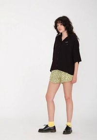 Chemise noire boutonnée à manches courtes, associée à un short jaune arborant un motif abstrait noir. Le mannequin porte des chaussures noires et des chaussettes jaunes.