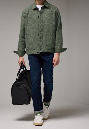 Mężczyzna w zielonej kurtce w wzory, ciemnoniebieskich dżinsach i białych trampkach, niosący w lewej ręce czarną torbę podróżną, idzie do przodu.
