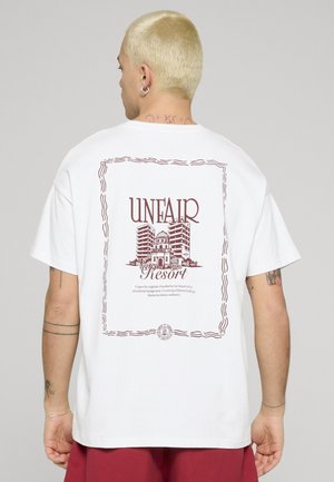 Person med kort blondt hår iført hvit t-skjorte som viser tekst og grafikk av bygninger på ryggen, innrammet med "UNFAIR Resort" i mørkerødt.