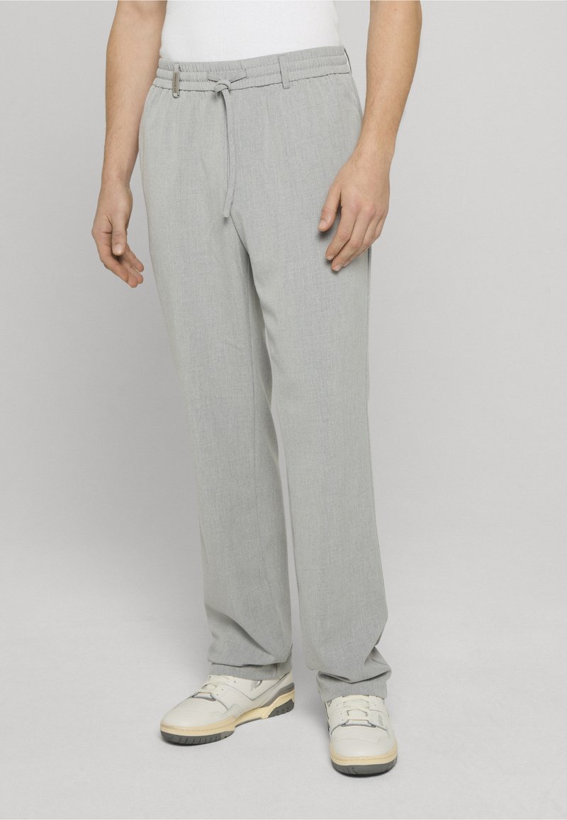Pantalon ample gris clair en tissu lisse avec taille élastique et cordon de serrage. Porté avec des baskets de couleur claire.