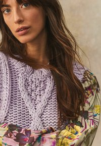 Pull en maille lavande avec un motif texturé en tresse ; écharpe à motifs floraux aux couleurs vives. Cheveux lisses sur un fond neutre.