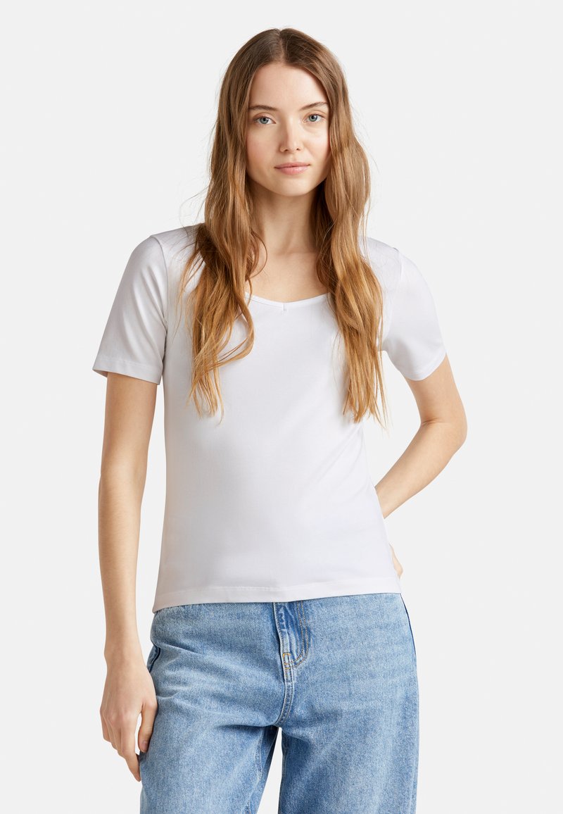 Mladá žena s dlouhými světlě hnědými vlasy, oblečená v bílém tričku s krátkým rukávem a modrých džínách, stojí s jednou rukou na bocích.