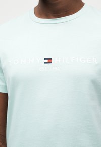 Ανοιχτό μπλε βαμβακερό t-shirt με στρογγυλή λαιμόκοψη. Φέρει κεντητό λευκό κείμενο "TOMMY HILFIGER" και ένα μικρό λογότυπο σε κόκκινο, άσπρο και μπλε.