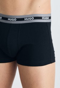 Schwarze Boxershorts mit einem grauen Bund, auf dem das Logo "HUGO" abgebildet ist. Der Stoff wirkt glatt, sitzt eng und hat minimale Nähte.