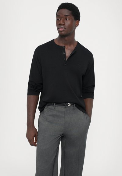 Čierna košeľa s dlhým rukávom a henley výstrihom, vybavená tromi gombíkmi. Nosená s svetlošedými, strihanými nohavicami a čiernym opaskom.