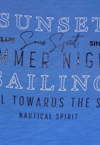 Camiseta azul con diseños de texto en diferentes fuentes y tamaños: "PUESTA DE SOL", "NAVEGACIÓN", "NOCHE DE VERANO", con un ligero brillo y textura suave.