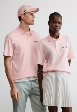 Δύο μοντέλα φορούν ανοιχτό ροζ πουκάμισα πόλο Lacoste· ο άνδρας με καπέλο και ανοιχτόχρωμο τζιν, η γυναίκα με πλισέ λευκή φούστα και σκουλαρίκια, στέκονται κοντά το ένα στο άλλο.