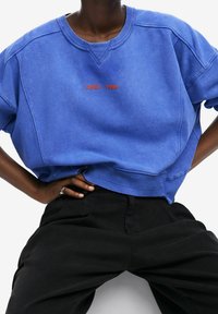 Felpa blu con scollatura rotonda e testo ricamato rosso "DSGL 1984." Presenta spalle scese e orlo corto. Abbinata a pantaloni neri.