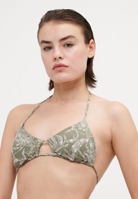 Tiskana bikini majica z dvema trikotnima deloma v olivno zeleni barvi z abstraktnimi belimi vzorci, nastavljivimi trakovi in podrobnostjo v obliki ključavnice na sredini.