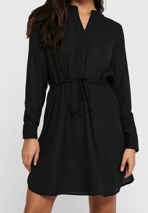 Vrouw die een zwarte blousejurk met lange mouwen en een taillekoord draagt, staand tegen een effen lichte achtergrond.