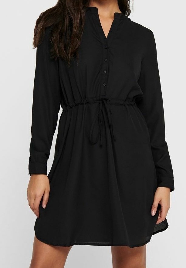 Femme portant une robe noire à manches longues boutonnée avec une ceinture à la taille, debout devant un fond clair uni.