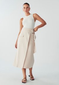 Ženska z belo brezrokavno majico, bež midi krilom z žepom in pasom, črnimi sandali z naramnicami, ki stoji pred enotno svetlo ozadjem.