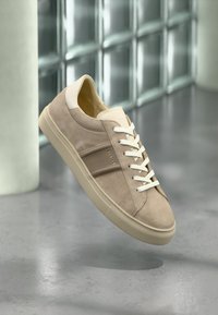 Μπεζ σουέντ αθλητικό παπούτσι με λευκά κορδόνια και λαστιχένια σόλα, αιωρείται πάνω από γκρίζο δάπεδο με φόντο τοίχο από γυάλινα τούβλα.