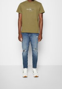 Olivgrön bomulls-T-shirt med en grå logotyp, sammansatt med ljusblå slitna jeans och vita sneakers med bruna detaljer.