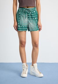 Pantaloni scurți verzi cu un model geometric, cu talie reglabilă prin șnur, accentuați cu detalii albe, purtați cu adidași albi cu guler înalt.