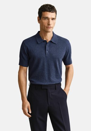 Mann trägt ein marineblaues Poloshirt mit kurzen Ärmeln, das in schwarze Hosen gesteckt ist, steht mit einer Hand in der Tasche vor weißem Hintergrund.
