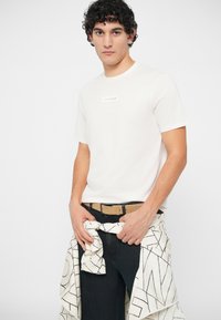 Man wearing white Armani Exchange t-shirt with black jeans, beige belt, and a patterned jacket tied around his waist against a white background.