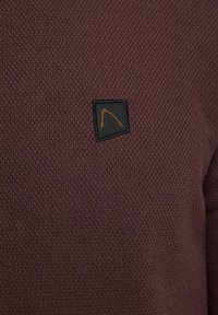 Pletena veste u tamnoj burgundiji s teksturiranim uzorkom. Ima malu crnu oznaku s izvezenim narančastim dizajnom.