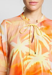 Lekka bluzka z tropikalnym nadrukiem palm w odcieniach pomarańczowym i żółtym. Posiada dekolt w kształcie łezki oraz żółty sznurek do troczenia przy szyi.