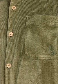 Chemise en velours côtelé vert olive avec nervures verticales, boutons en bois et une poche poitrine unique ornée d'un logo discret.