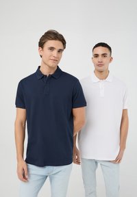 Granatowe i białe koszulki polo wykonane z mieszanki bawełny, z klasycznymi kołnierzykami, krótkimi rękawami i zapięciem na guziki na neutralnym tle.